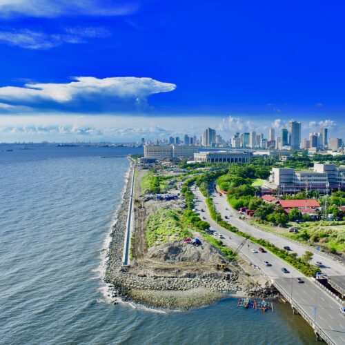Aerial image of the Manila skyline from the seaside. The left side of the image is open water, and the right side starts with a rocky coast and road with some tall buildings in the background.