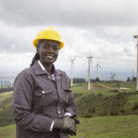 A woman in a yellow hardhat stands on a hill near several wind turbines, smiling at the camera.