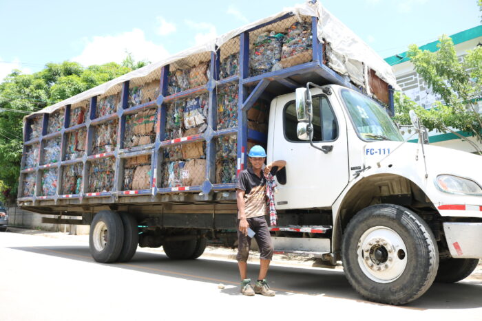 A man in a blue work helmet stands in front of a large trailer truck filled to the brim with bald plastic recyclables.