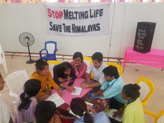 A group of men and women sit together at a table, talking and studying. Behind them, a sign says "Stop Melting Life, Save the Himalayas."