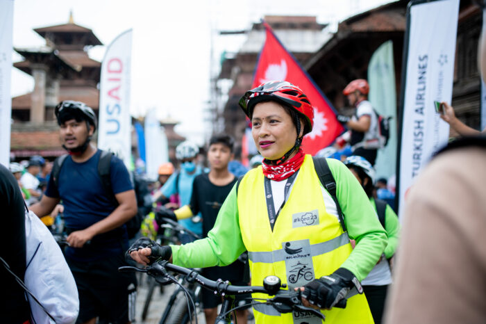 A woman in a high-visibility vest and a red bicycle helmet walks toward the camera smiling and holding her bicycle handles. 