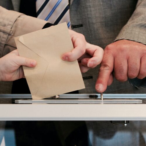 Image: Three hands hover over a voting box, while two of the three hands grasp an envelope as it is placed into a slot on top of the box.
