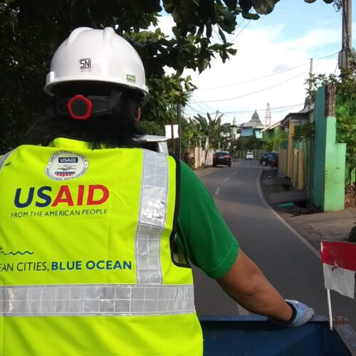 A man wearing a high visibility vest with the USAID logo on the back is facing away from the camera.