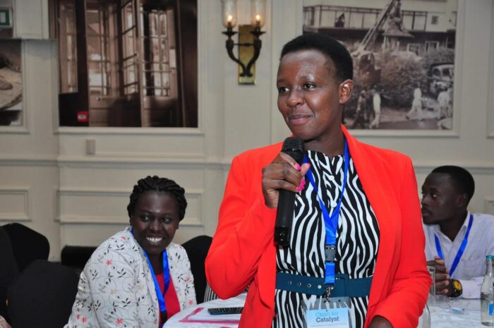 A woman stands holding a microphone, speaking to people at a work shop.