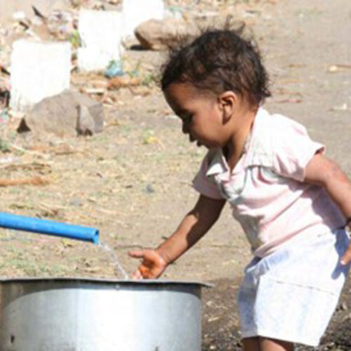 A young child places her hand into water flowing from a blue hose into a metallic vessel.