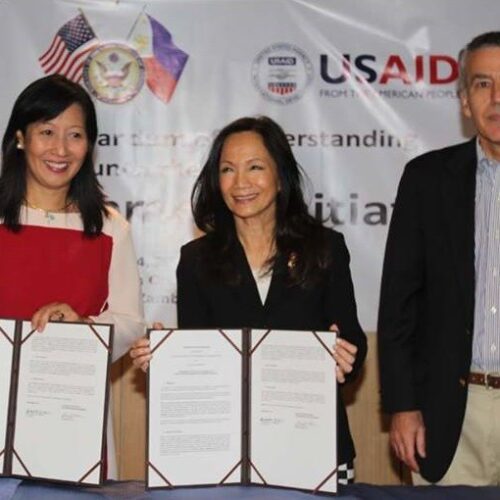 One man and two women stand smiling in front of a banner with US and Philippine flags alongside the USAID logo.