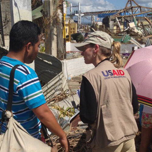 A man and a woman wearing a vest with the USAID logo on the back look at a disaster scene.