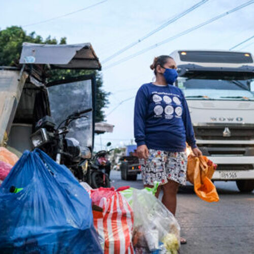 A woman wearing a mask stands next to several full plastic bags on the side of a road as a large truck passes.
