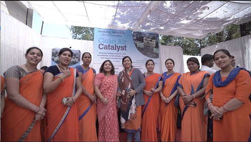 A group of women in orange sari pose for the camera in front of a Clean Air Catalyst sign.