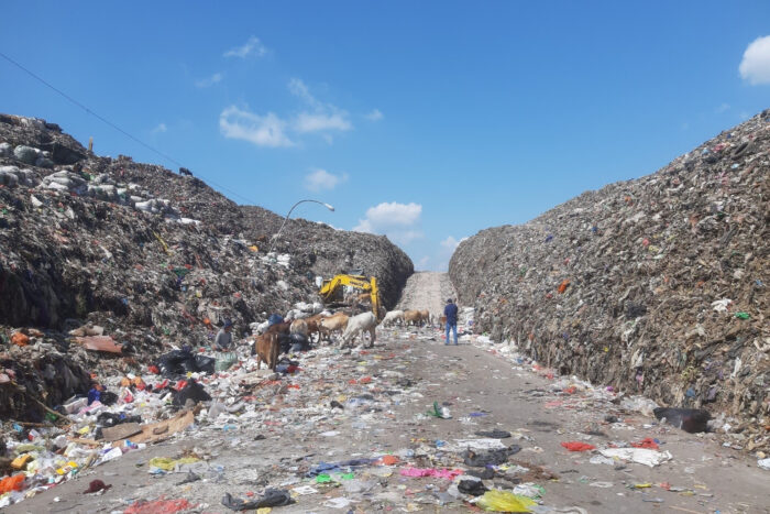 Street view of a dump from the interior, with high unmanaged piles of trash lining each side of a street that is center of frame.