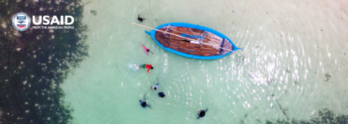 Photo showing aerial view of medium boat floating in clear waters off coast of the Maldives with several people swimming around the boat.