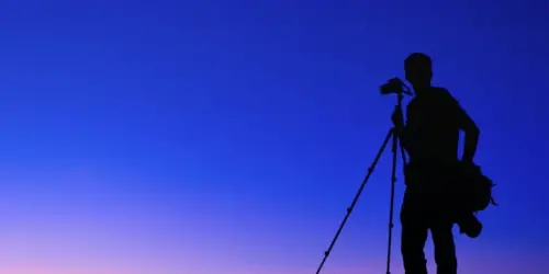 Silhouette against a blue gradient background of a man with photo gear using a camera on a tripod.