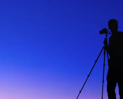 Silhouette against a blue gradient background of a man with photo gear using a camera on a tripod.