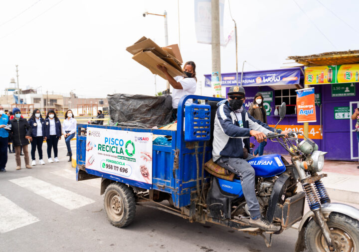 A man drives a motorcycle that has been converted into a trailer, while a woman stands in the trailer section and loads cardboard.