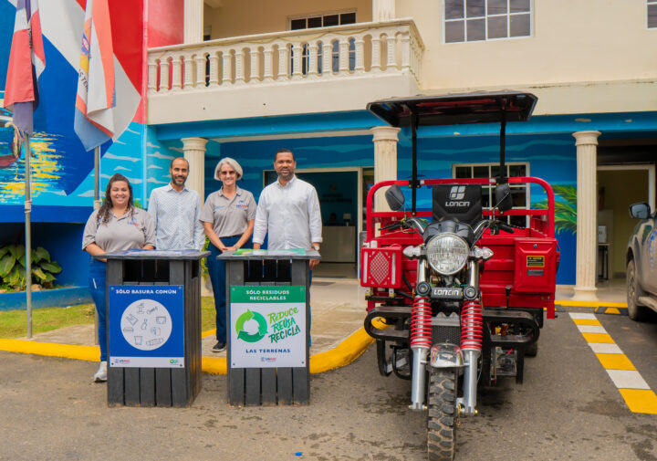 Two couples pose for the camera, standing behind waste collection bins and next to a large red motorized tricycle.