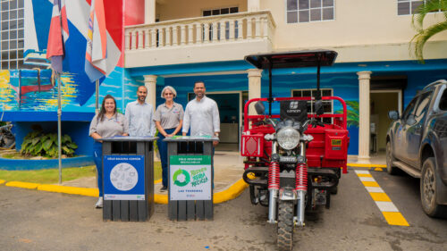 Two couples pose for the camera, standing behind waste collection bins and next to a large red motorized tricycle.