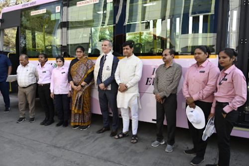 Several men and women, some in pink and black uniforms, stand beside a pink bus and pose for a group photograph.