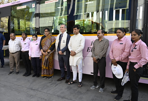 Several men and women, some in pink and black uniforms, stand beside a pink bus and pose for a group photograph.