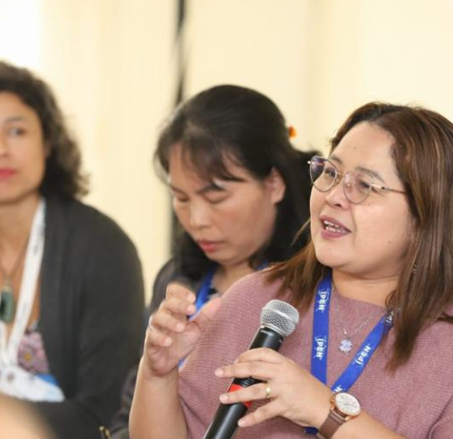 A woman smiles while speaking into a microphone in a meeting setting. In the background, another woman takes notes, and another looks at the speaker.