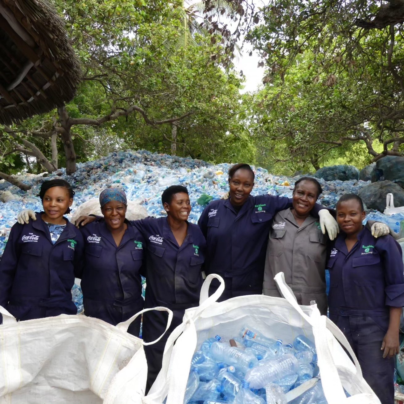 A group of five women in uniform stand smiling for the camera in front of a large pile of plastic bottles.