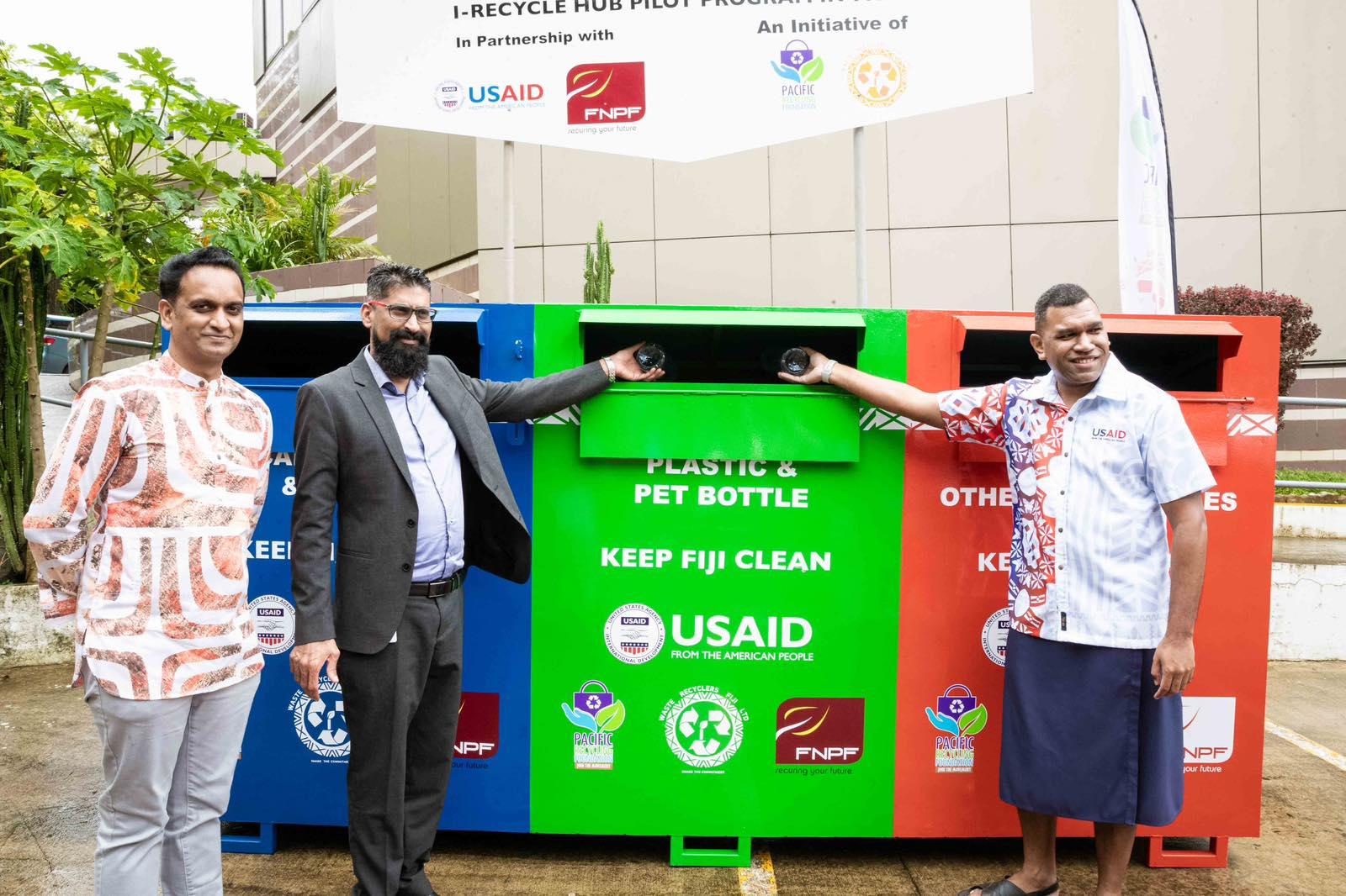 Three men pose in front of multicolored recycling bins, smiling as they drop glass bottles into a central green bin.