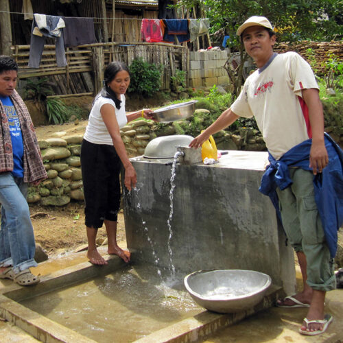 Two men and a woman stand near a water source as water flows from a spigot into a drain area.