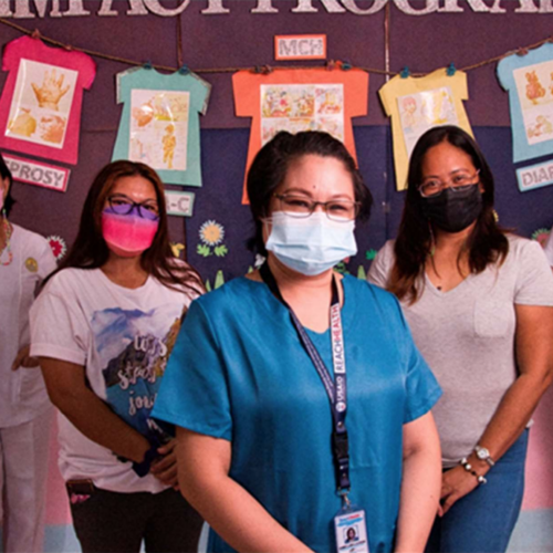 Five women wearing masks stand in front of a colorful background, looking at the camera.