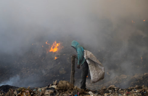 a man stands in a landfill, where there is an open flame in the background.