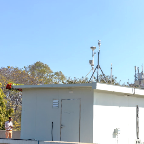 A man stands next to a small white building with multiple antenna and other monitoring apparatuses on top of it.
