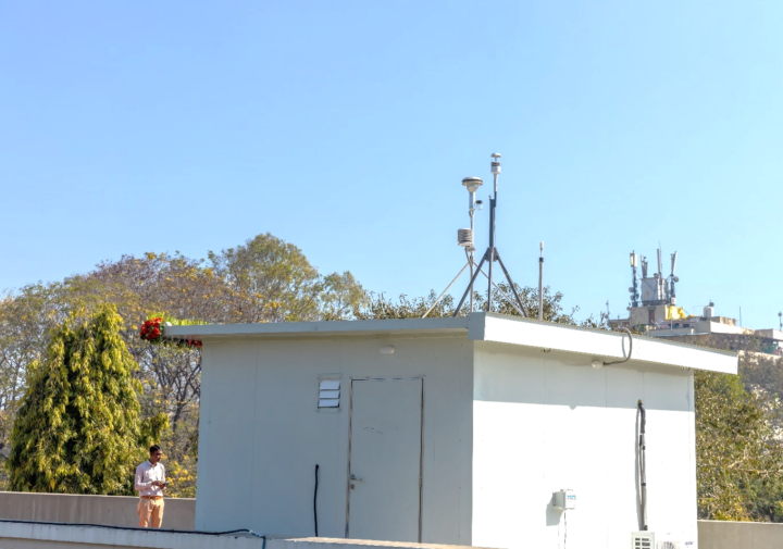 A man stands next to a small white building with multiple antenna and other monitoring apparatuses on top of it.