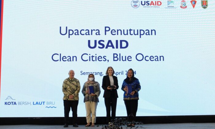 One man and three women pose in front of a large screen that reads "USAID Clean Cities, Blue Ocean" in English and Bahasa Indonesia.