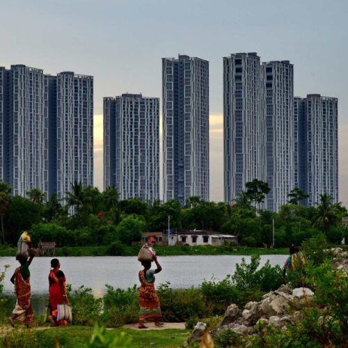 Three women walk along a riverbank. In the background, several high-rise apartment buildings create a contrast with the greenery and low buildings in the foreground.