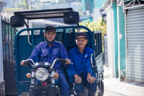 A Vietnamese man and woman in matching blue uniforms smile at the camera while driving a motorcycle with attached trailer.