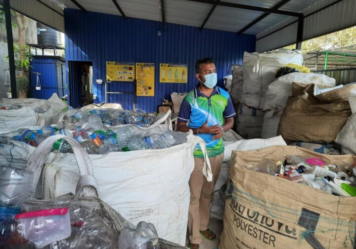 A man stands in an open warehouse among large bags filled with plastic bottles.
