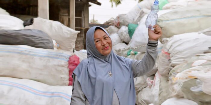 Woman standing in front of large mounds of gathered trash holding up a plastic bottle.